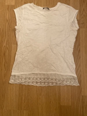 Vit blus med spetskant från Lindex - Säljer en vit blus från Lindex i storlek 170. Toppen har korta ärmar, rund hals och en liten bröstficka med spetsdetalj. Nederkanten är dekorerad med bred spets med blommönster som ger en söt touch. Perfekt för dig som gillar romantisk stil.