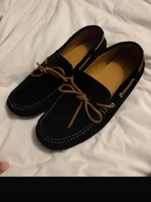 Svarta mocka loafers med snörning - Snygga svarta loafers i mocka med bruna snören och vita sömmar. Skorna har en klassisk seglarsko-design med rund tå och platt sula. Perfekta för dig som gillar stilrena och tidlösa skor med en chill vibe.