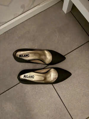 Svarta pumps med spetsig tå från Milanc - Svarta pumps från Milanc i storlek 37 med elegant spetsig tå och låg klack. Skorna har en stilren design med insida i guldton och är tillverkade i ett syntetmaterial som ger en matt finish. Perfekt för dig som gillar klassisk och chic stil.