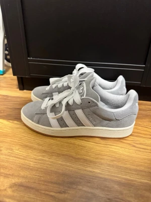 Adidas Campus  - Snygga Adidas sneakers i grå mocka med klassiska vita ränder på sidorna och vit logga på plösen. Skorna har vit snörning och en ljusbrun gummisula med mönster. Skorna är använda 1-2 gånger, som man ser på bilden.