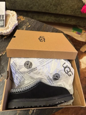 Svarta UGG Dakota mockatofflor - Säljer ett par svarta UGG Dakota tofflor i mocka med fluffigt foder och mönstrad kant i vitt och grått. Snygg rund tå och platt sula med grovt mönster undertill. Perfekt för chill dagar hemma eller när du vill ha det extra bekvämt.