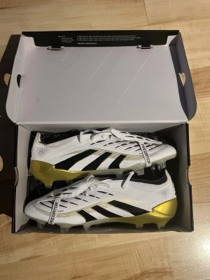 Adidas Predator fotbollsskor vit/guld - Säljer ett par Adidas Predator fotbollsskor i vitt med svarta och guldiga detaljer. Skorna har snörning, låg profil och platt sula med dobbar för gräsplan. Materialet är syntet och designen har klassiska svarta ränder på sidan samt guld på hälen och yttersulan.