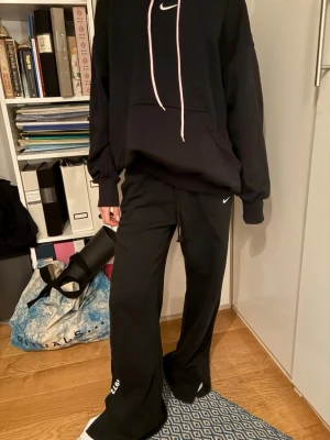 Svarta Nike mjukisbyxor wide fit - Svarta mjukisbyxor från Nike med bred passform och dragsko i midjan. Byxorna har vit Nike-logga framtill och tryck med text och årtalet 1972 på ena benet. Sköna och avslappnade, perfekta för chill eller träning.