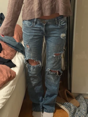 Blå slitna jeans från Lee - Säljer ett par blå jeans från Lee med flera stora slitningar och hål på benen för en riktigt edgy vibe. Modellen har klassisk femficksdesign, låg midja och raka ben. Jeansen är gjorda i robust bomullstyg och har Lee-logga bak på linningen.