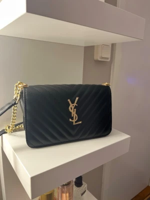 Svart quiltad axelväska från YSL - Säljer en svart quiltad axelväska från YSL med guldfärgad kedjerem och ikonisk YSL-logga framtill. Väskan är i skinn och har ett rymligt fack med dragkedja inuti. Perfekt storlek för det viktigaste och ger en lyxig vibe till din outfit.