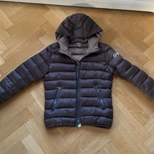 Mörkbrun Colmar pufferjacka med huva - Snygg mörkbrun pufferjacka från Colmar med huva och två fickor med dragkedja. Jackan har ett quiltat mönster och logotyp på ärmen. Perfekt för kalla dagar och har en stilren look som funkar till många outfits.