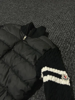 Svart pufferjacka från Moncler - Svart pufferjacka från Moncler med stickade ärmar och vita ränder på armarna. Jackan har dragkedja framtill, två sidofickor med dragkedja och ribbad krage. Moncler-logga på vänster ärm. Perfekt för dig som vill ha en unik jacka med coola detaljer.