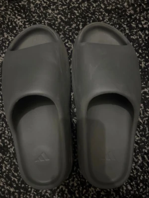 Grå Adidas slides med räfflad sula - Säljer ett par grå Adidas slides med minimalistisk design och räfflad sula för extra grepp. Skorna är tillverkade i syntetmaterial och har öppen tå samt bred rem över foten. Haft i två månader. Storlek 46