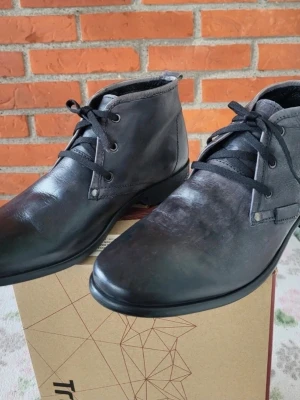 Mörkgråa skinnkängor med snörning från Legend 41 - Snygga smokgråa kängor från Legend i äkta skinn med klassisk snörning och rund tå. Skorna har en stilren design med låg profil och platt sula, perfekta för dig som gillar enkel och tidlös stil. Passar till jeans eller chinos och funkar året runt.