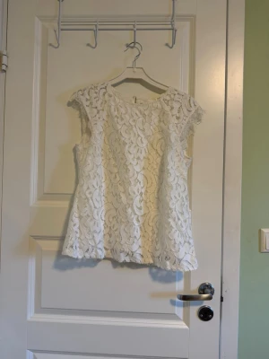 Vit spetsblus - Säljer en vit blus med vackert blommönstrad spets. Blusen är ärmlös och har rund halsringning. Passar till både jeans och kjol för en snygg look.