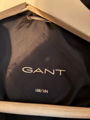 Mörkblå pufferjacka från GANT - Snygg mörkblå pufferjacka från GANT med huva och dragkedja framtill. Jackan har quiltad design och långa ärmar, perfekt för kalla dagar. GANT-logga syns på kragen och jackan har praktiska fickor på sidorna.