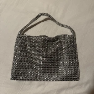 Glittrig silver handväska - Säljer denna glittriga handväskan i silver med massor av små stenar som ger ett festligt intryck. Väskan har ett kort handtag. Den är helt oanvänd & i fint skick!💕