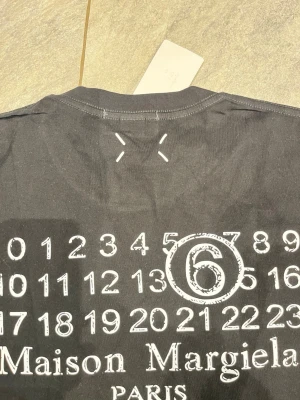 Maison Margiela svart t-shirt med print - Svart t-shirt från Maison Margiela med ikoniskt siffror-print och cirkel runt nummer 6. Klassisk rund hals och korta ärmar. T-shirten har vita stygn på ryggen och är gjord i mjuk bomull. Perfekt för dig som gillar statement-plagg och cool design.