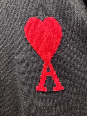 Svart stickad tröja från Ami - Svart stickad tröja från Ami Alexandre Mattiussi med ikonisk röd hjärt-A broderad på bröstet. Tröjan har rund hals, ribbade muddar och en loose passform som ger en chill vibe. Perfekt för dig som gillar stilrena detaljer med en twist.