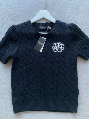 Svart kabelstickad tröja Lauren Ralph Lauren - Kabelstickad svart tröja från Lauren Ralph Lauren med korta puffärmar och vit broderad RL-logga på bröstet. Klassisk rund halsringning och ribbade muddar. Aldrig använd!💕