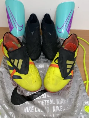 Pris kan diskuteras Adidas Predator fotbollsskor  två par - Två par Adidas Predator fotbollsskor, ett par i svart med silvriga detaljer och ett par i gul med svarta och orange detaljer. Dess par som är gula har täckt snörning och mönstrad sula för extra grepp på planen. Däremot det svarta paret av fotbollskor har ingen snörning och passar som en snrumpa, även detta par är perfekt för dig som vill dominera på planen. Båda är  Perfekta för dig som vill sticka ut på planen. Paren som är gula har storlek 40 däremot är det andra paret storlek 42,2/3.