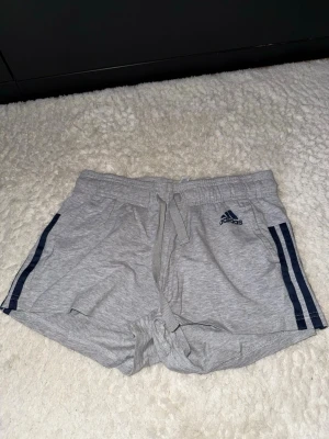 Grå Adidas shorts med snörning - Säljer ett par gråa Adidas shorts med marinblå ränder på sidorna och logga framtill. De har snörning i midjan och är gjorda i mjuk bomull, perfekta för chill eller träning. Passformen är normal och de är korta i modellen.