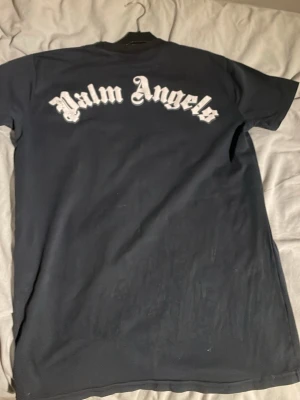 Svart Palm Angels t-shirt med tryck - Svart t-shirt från Palm Angels med klassiskt tryck i vitt på ryggen och logga broderad vid halsen. Kortärmad modell med rund hals och extra Palm Angels-detalj vid nederkanten. Skön bomullskvalitet och clean streetwear-vibe.