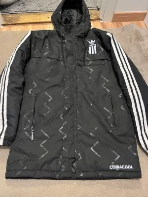 Svart Adidas vindjacka med mönster - Säljer en svart Adidas vindjacka med huva och coola silvriga zigzag-mönster. Jackan har vita ränder längs ärmarna, flera dragkedjeförsedda fickor och Climacool-tryck nedtill. Perfekt för dig som vill ha sportig stil och funktion i ett.