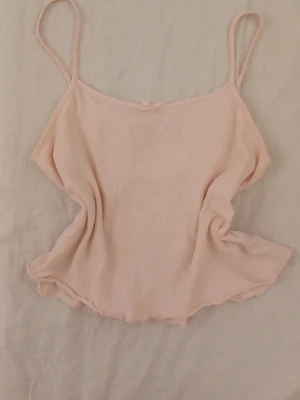 Ljusrosa linnetopp med rosett - Supersöt ljusrosa linnetopp med tunna axelband och en liten rosett framtill. Toppen har en lätt vågig kant nedtill och är tillverkad i ett mjukt, ribbat material som känns skönt mot huden. Perfekt för varma dagar och enkel att matcha med jeans eller kjol.