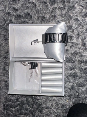 Svart Lotus klocka med metallarmband - Snygg svart Lotus klocka med stilrent metallarmband och klassisk rund urtavla. Urtavlan är svart med vita detaljer och datumvisning. Kommer i original ask med extra länkar. Perfekt accessoar för dig som gillar en clean och modern look. Har några få större skrap repor som syns - bild bifogad!  Säljs för att min kille köpt ny           Nypris på klockan är - 1200 kr                    Jag säljer denna för 600 kr men priset är inte hugget i sten! 