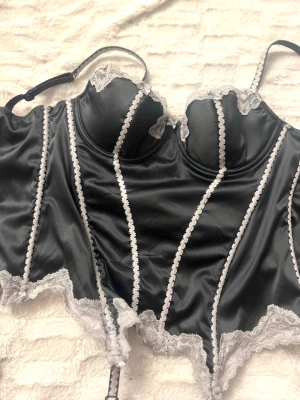 Svart satinväst med spets och rosetter - Supersnygg svart korsett i satin med vita spetsdetaljer och små rosetter. Västen har reglerbara axelband och dekorativa vertikala spetsband framtill. Spetsen pryder både kuporna och nederkanten för en extra lyxig vibe. Perfekt för dig som gillar statement-plagg med lite edge. Passar storlek XS / S