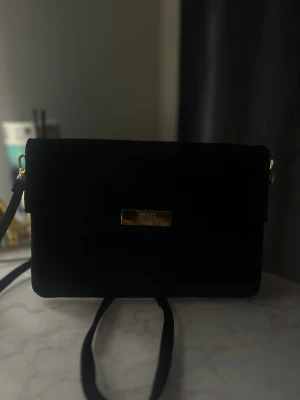 Svart mocka axelväska från BOSS - Stilren svart axelväska från BOSS i mjuk mocka med rektangulär form och guldfärgad metallplatta framtill. Väskan har en smal axelrem och minimalistisk design som ger en lyxig vibe. Perfekt för dig som gillar klassiska accessoarer med en modern touch.