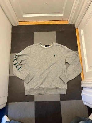 Grå sweatshirt från Polo Ralph Lauren - Snygg unik grå sweatshirt från Polo Ralph Lauren i bomullsmix. Klassisk rund hals, långärmad med grönt Polo-tryck på ena ärmen och liten grön logga på bröstet. Perfekt för chill dagar och enkel att matcha med jeans eller joggers.