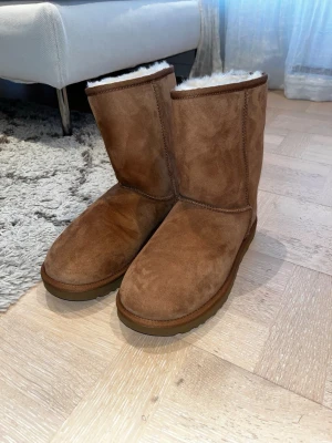 Bruna UGG boots i mocka - Säljer ett par klassiska UGG boots i brun mocka med rund tå och platt sula. Insidan är fodrad med mjuk, ljus päls som håller fötterna varma. Perfekta för kyliga dagar och har en enkel, tidlös design med UGG-logga bak på hälen. Storlek 41, aldirg använda, ser helt nya ut