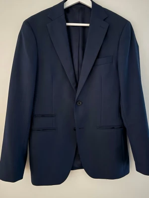 Marinblå kavaj med klassisk passform - Snygg marinblå kavaj från these glory days med klassisk design och två knappar framtill. Kavajen har smala slag, tre framfickor och knappar vid ärmsluten. Materialet är slätt och känns lyxigt, perfekt för dig som vill ha en stilren look. Passar dig som gillar enkel och tidlös stil.