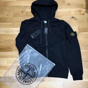 Stone Island Zip Hoodie - Black Stone Island Zip Hoodie Stilren och bekväm zip-hoodie från Stone Island i klassisk svart färg. Mjuk, varm och perfekt för vardag eller casual outfits. En tidlös hoodie som kombinerar stil och funktion – perfekt för vintern och kyliga dagar. Även har QR-CODE. TVEKA INTE ATT SKICKA EN BUD