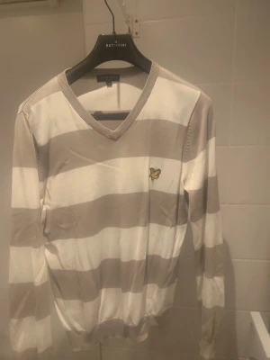 Randig v-ringad tröja från Lyle & Scott - Snygg v-ringad tröja från Lyle & Scott med breda beige och vita ränder. Tröjan har lång ärm och ett broderat guldfärgat fågelmärke på bröstet. Mjuk och tunn i bomull, perfekt för dig som gillar klassisk stil med en modern touch.