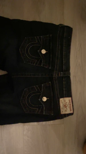 True Religion mörkblå bootcut jeans - Säljer ett par mörkblå true religion jeans som e i princip oanvända från True Religion med bootcut passform. Jeansen har markerade kontrastsömmar, stora fickor med lock och metallknappar med logga. Klassisk patch bak i midjan och detaljerad brodering på fickorna.  De är lite lätt utslitna precis längst ner.