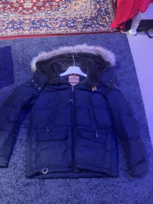 Mörkblå dunjacka Parajumpers med pälskrage - Säljer en mörkblå dunjacka från Parajumpers med fluffig pälskant på huvan. Jackan har flera fickor med dragkedja och knappar, samt detaljer som ribbade muddar och justerbara ärmslut. Perfekt för kalla vinterdagar och riktigt snygg streetstil. Har lite små håll