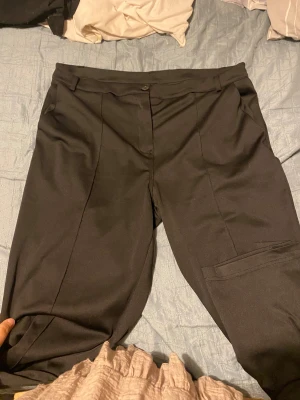  kostymbyxor med pressveck - Snygga svarta kostymbyxor med klassiska pressveck och bälteshällor. Byxorna har rak passform och är tillverkade i ett slätt material som ger en stilren look. Perfekta för dig som vill ha en clean och enkel stil. Stängs med knapp och dragkedja framtill.