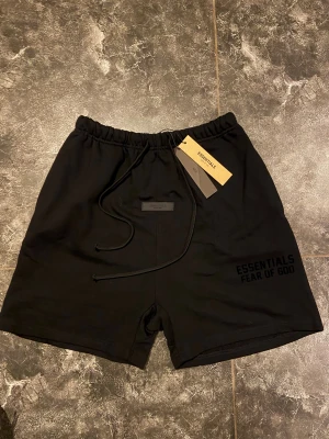 Svarta shorts Essentials Fear of God - Pris kan diskuteras. Bra skick. Svarta shorts från Essentials Fear of God med stilren logga framtill. Modellen har elastisk midja med snörning och en bakficka. Materialet är mjuk bomull som känns skönt mot huden. Perfekt för dig som gillar streetwear och vill ha en clean look.