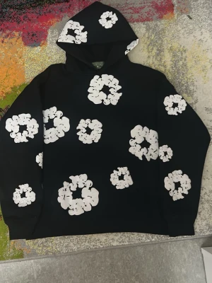 Svart och vit hoodie från denim tears - Stilren hoodie i svart med unikt, handmålat-inspirerat mönster av vita blom-/molnliknande cirklar över hela plagget. Bekväm passform med huva och magficka. Bra för alla väder