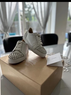Vita sneakers med nitar från Louboutin - Säljer ett par vita sneakers från Christian Louboutin med coola silverfärgade nitar på tå och häl. Skorna har snörning och är tillverkade i skinn med platt sula och rund tå. Perfekt för dig som vill sticka ut med en edgy och lyxig stil.
