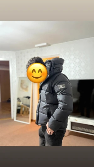 Svart Burberry pufferjacka med huva - Säljer en svart pufferjacka från Burberry London England med stor, mysig huva och snörning. Jackan har tydliga quiltade sektioner, dragkedja och knappar framtill samt logotyp på ärmen. Perfekt för kalla dagar och riktigt snygg streetstil.