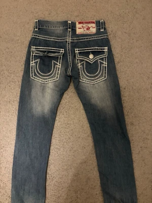 Vintage True Religion jeans - Säljer ett par blå vintage True Religion Joey Super T jeans med kontrasterande vit söm och stora fickor med lock och knapp bak. Klassisk bootcut-modell med låg midja och slitningar för cool vintagekänsla. Obs!! Saknar en knapp!💘