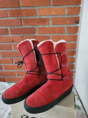 Röda mockaboots från Catwalk Stockholm - Säljer ett par röda boots från Catwalk Stockholm med svart platt sula och snörning runt skaftet. Utsidan är i mocka och insidan är fodrad med mjuk, vit pälsimitation. Snygga detaljer med röda remmar och rund tå. Perfekta för kalla dagar!