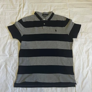 Randig pikétröja från Polo Ralph Lauren - Säljer en snygg pikétröja från Polo Ralph Lauren i custom slim fit strl M/L. Tröjan har breda horisontella ränder i mörkblått och grått, klassisk krage med två knappar och korta ärmar. Ikoniska Polo-loggan broderad på bröstet. Perfekt för en chill och stilren look.