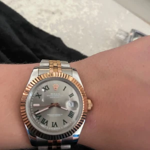 Rolle Datejust tvåtonad klocka - Säljer en stilren Rolle Datejust med tvåtonad länk i silver och guld. Klockan har en rund boett, räfflad guldfärgad bezel och silverfärgad urtavla med romerska siffror. Länken är i stål och guld, och kronan samt detaljer är i guld. Klassisk och tidlös design.