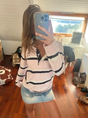 Randig rosa henley från GANT - Snygg långärmad henley från GANT i ljusrosa med breda mörkblå och vita ränder. Skjortan har klassisk krage, knappar i halsen och är gjord i mjuk bomull. Perfekt för dig som gillar klassik stil och vill ha något som sticker ut lite extra.