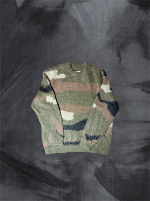 Camouflage stickad tröja i ull - Säljer en asfet stickad tröja med camouflage-mönster. Tröjan har rund hals och långa ärmar, perfekt för dig som gillar streetwear och vill ha en unik look. Materialet är mjuk ull som känns skönt mot huden. 