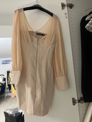 Champagnefärgad kort klänning Lavish Alice - Snygg kort klänning från Lavish Alice i champagnefärg med transparenta långärmar och breda manschetter. Klänningen har en djup v-ringning och markerad midja som ger en elegant siluett. Perfekt för dig som vill sticka ut med stil.