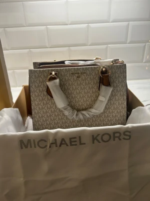 Helt ny Beige handväska från Michael Kors  - Snygg handväska från Michael Kors i beige med MK-logga över hela väskan. Väskan har bruna detaljer, guldfärgade metalldelar och dragkedja upptill. Stilren och boxig form med dubbla handtag. Perfekt för dig som gillar klassisk design med lyxig touch. Aldrig använd helt ny med kvitto, box, påse och prislapp på. Nypris 3900kr mitt pris 1500kr