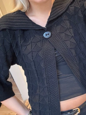 Svart stickad kofta med stor krage - Säljer en svart stickad kofta med snygg struktur och stor krage. Koftan har trekvartsärmar och stängs med en stor knapp framtill. Perfekt för lager på lager och ger en cool, avslappnad vibe till din outfit.