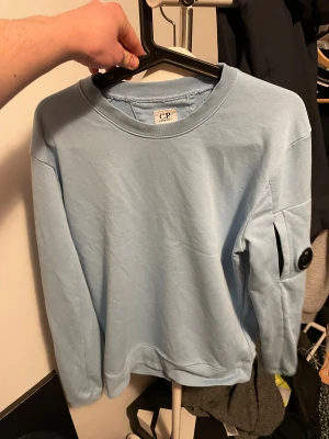 Ljusblå sweatshirt från C.P. Company - Snygg ljusblå sweatshirt från C.P. Company med klassisk rund hals och långärmad passform. Tröjan har en ikonisk svart linsdetalj på ärmen som ger en unik streetwear-vibe. Tillverkad i mjuk bomull för skön komfort och stil.