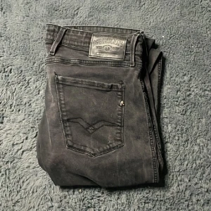 Svarta Replay Anbass jeans W34 L30 - Svarta Replay Anbass jeans i klassisk femficksmodell med raka ben och snygga detaljer på bakfickorna. Jeansen har normal passform och är tillverkade i mjukt bomullsmaterial med lite stretch för extra komfort. Perfekta för dig som gillar stilrena svarta jeans. 